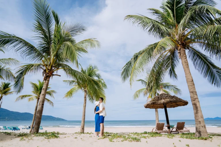 A young couple embracing on a tropical beach surrounded by palm trees under a bright sky.
