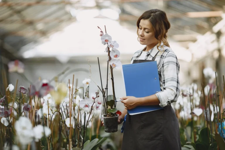 Woman in greenhouse holding a potted orchid and clipboard, surrounded by flowers.